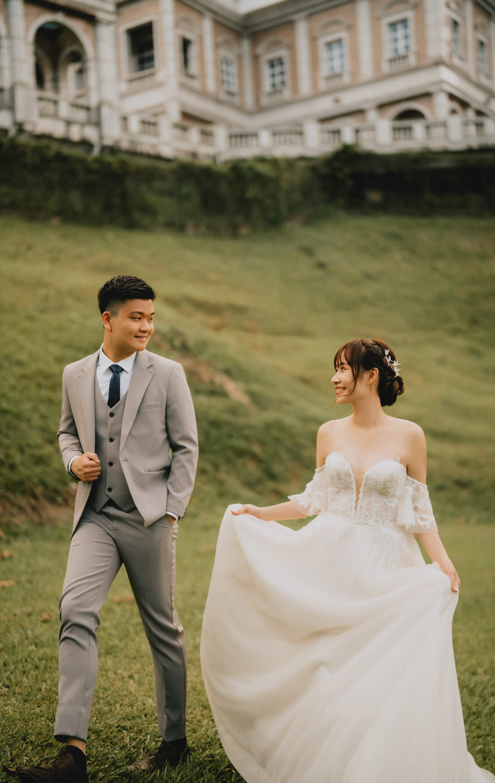A bride and groom stroll hand in hand across the grass, with a grand mansion in the background.